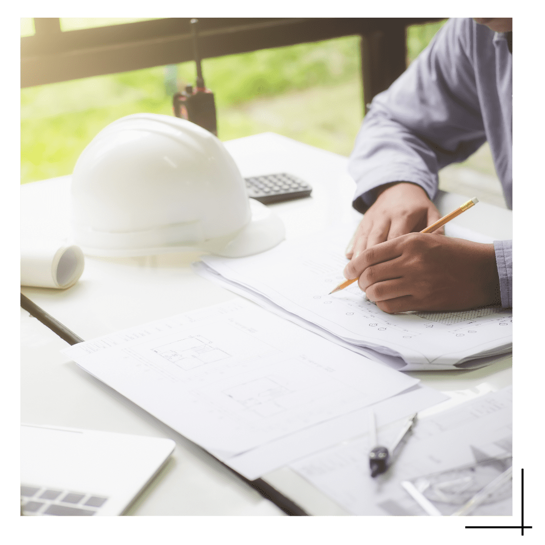 foto de um engenheiro sentado em uma mesa escrevendo em um papel, é possível ver apenas suas mãos e parte do seu terno, ao seu lado está um chapéu de obra na cor branca, representando que ele é um engenheiro.